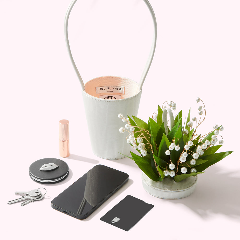 A ivory white Lulu Guinness bucket bag sits next to a small arrangement of lily of the valley, a smartphone, keys, a compact mirror, and a credit card, all on a white background.