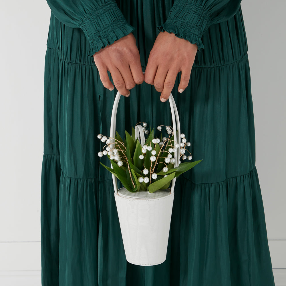 A person in a green dress holds a white bucket bag decorated with pearl lilies.
