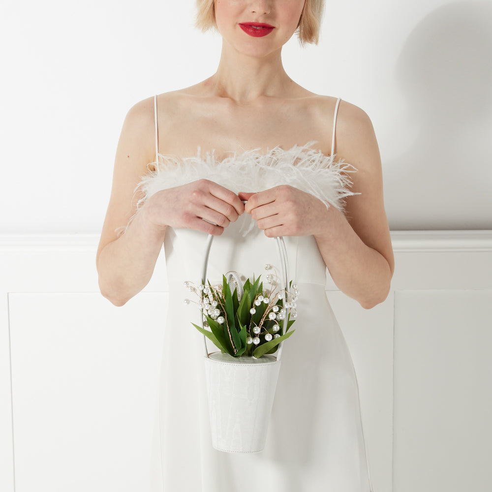 Woman in white dress holding a white handbag decorated with lily of the valley and pearls. The background is white.
