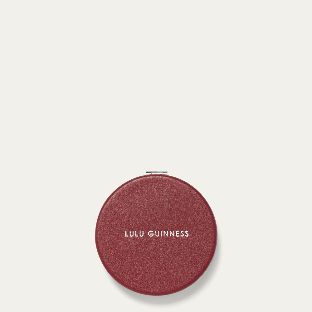 Round burgundy red compact mirror with 'Lulu Guinness' branding on a light beige background
