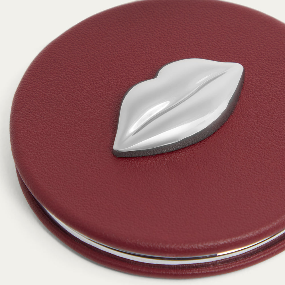 Burgundy red leather compact mirror with a silver lip-shaped emblem on a white background