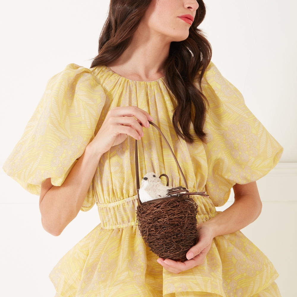 A close up image of a model with dark hair and red lipstick wearing a yellow occasion dress and holding a bird nest shaped bag.
