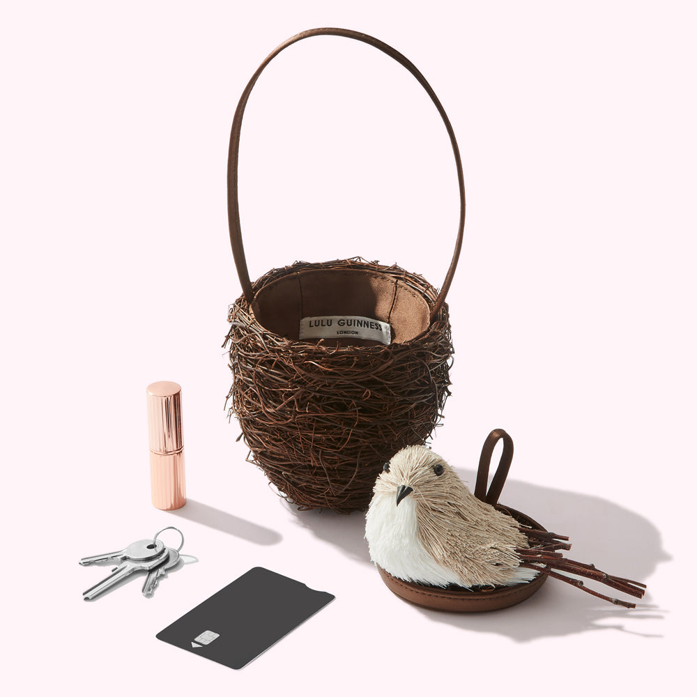 A brown bird's nest-shaped handbag with a small bird charm sits on a white surface next to keys, a credit card, and lipstick.