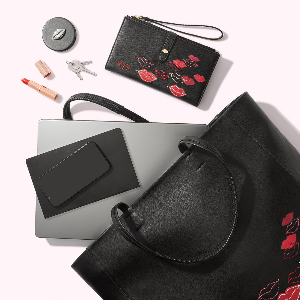 A black tote bag with red lips embroidered on the side, a laptop, a black wallet with red lips, lipstick, keys, and a compact mirror are arranged on a white background.
