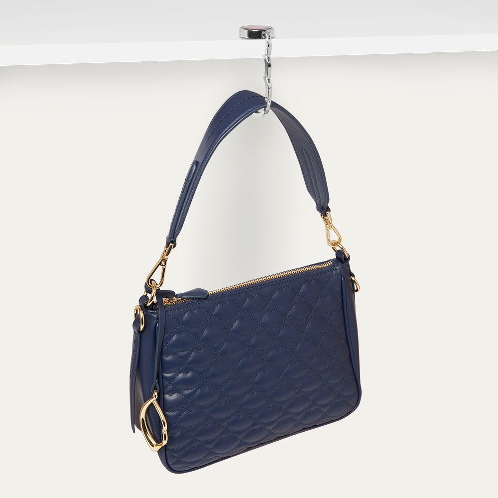 Navy blue handbag with gold accents hanging on a silver bag hook on a white background