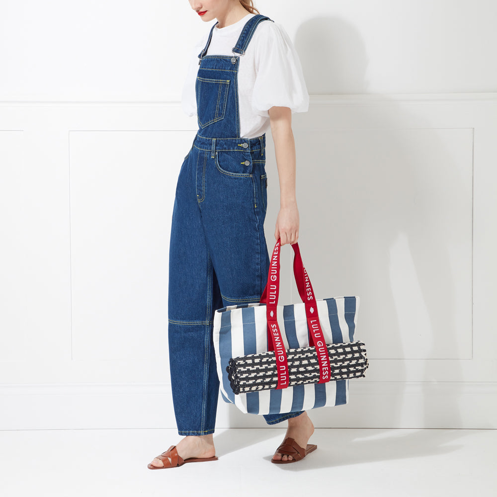 Woman in denim overalls holding a striped Lulu Guinness tote bag against a white background.
