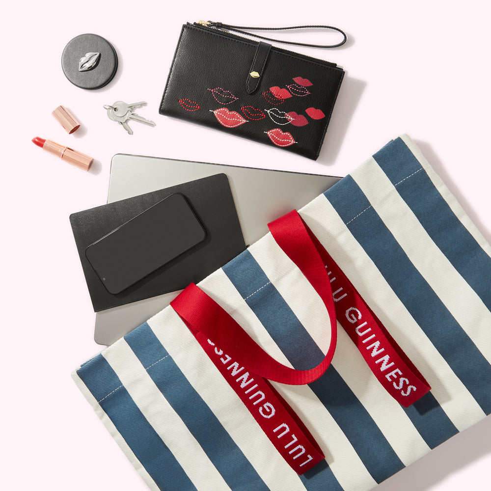 Flatlay of a Lulu Guinness striped tote bag, a black lip-print wallet, laptop, phone, lipstick, compact mirror and keys on a white background.