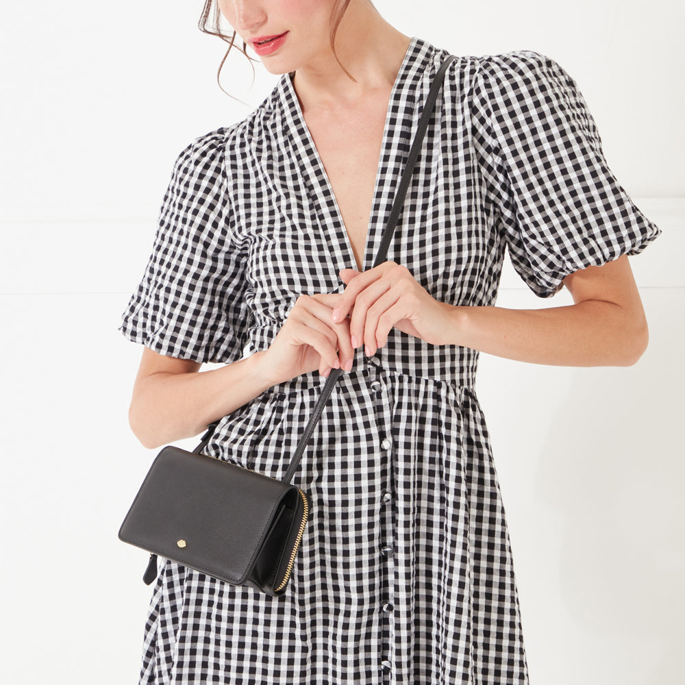 A close-up image of a model with dark hair wearing a black and white gingham dress and a black leather crossbody bag with a gold lip shaped pin on the front.