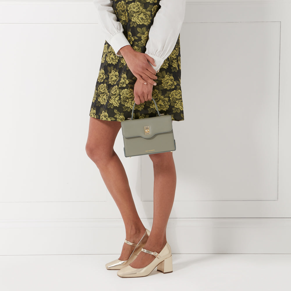 Woman in floral skirt holding a sage green handbag and wearing gold shoes against a white background.
