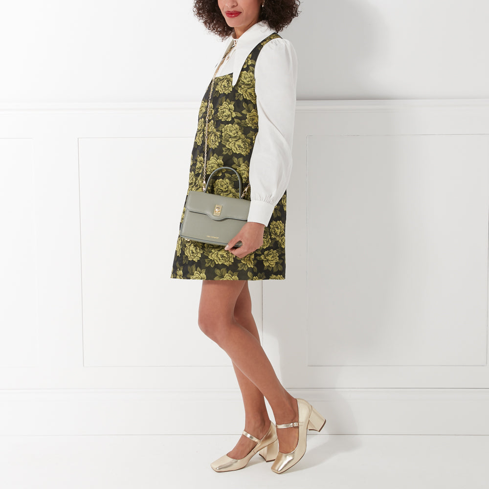 A woman in a floral dress and gold shoes carries a sage green handbag.