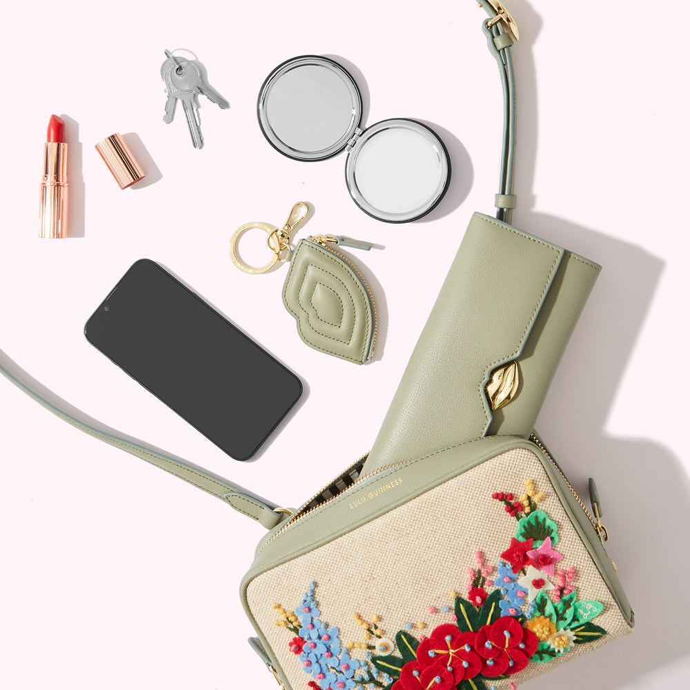 A flatlay of a LULU GUINNESS crossbody bag with embroidered flowers, a sage green wallet, compact mirror, lipstick, keys, and smartphone. The bag is open, displaying its interior.
