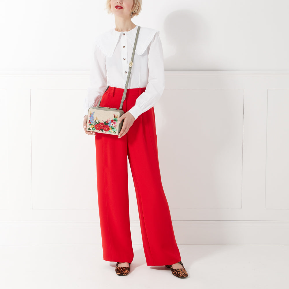 A woman in a white blouse and red pants holds a light beige crossbody bag with embroidered flowers. The bag has a sage green strap. The background is white.

