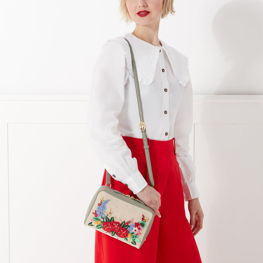 Woman in white shirt and red pants holding a beige crossbody bag with embroidered flowers.