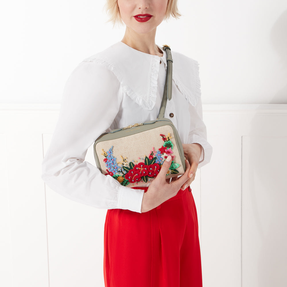 Woman in white shirt and red pants holding a sage green crossbody bag with floral embroidery.

