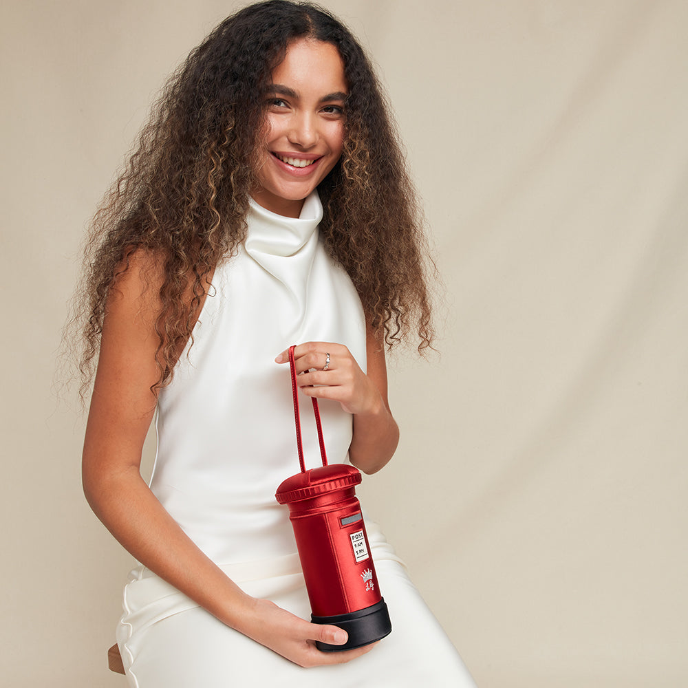 Woman holding a red satin postbox shaped handbag against a beige background