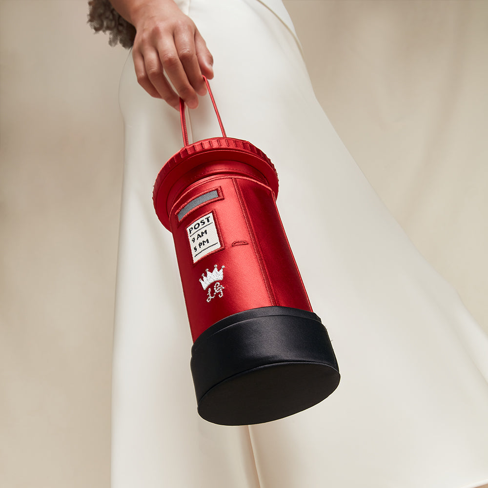Red satin postbox shaped handbag with black base held by a person against a neutral background