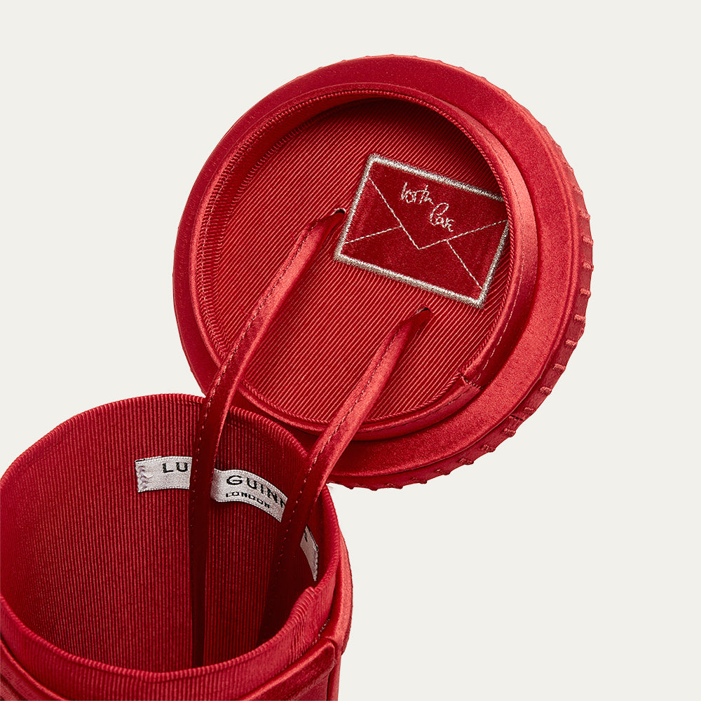 Close-up image of a red satin postbox shaped handbag with the lid open to reveal embroidered detail on a light gray background