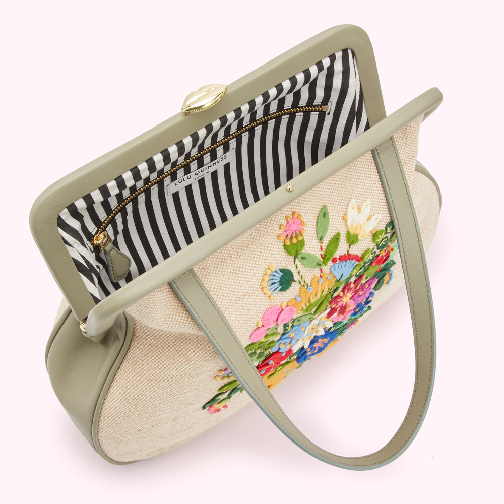 Overhead view of a Lulu Guinness cream handbag with sage green leather accents and a colourful embroidered floral bouquet. The interior is black and white striped.
