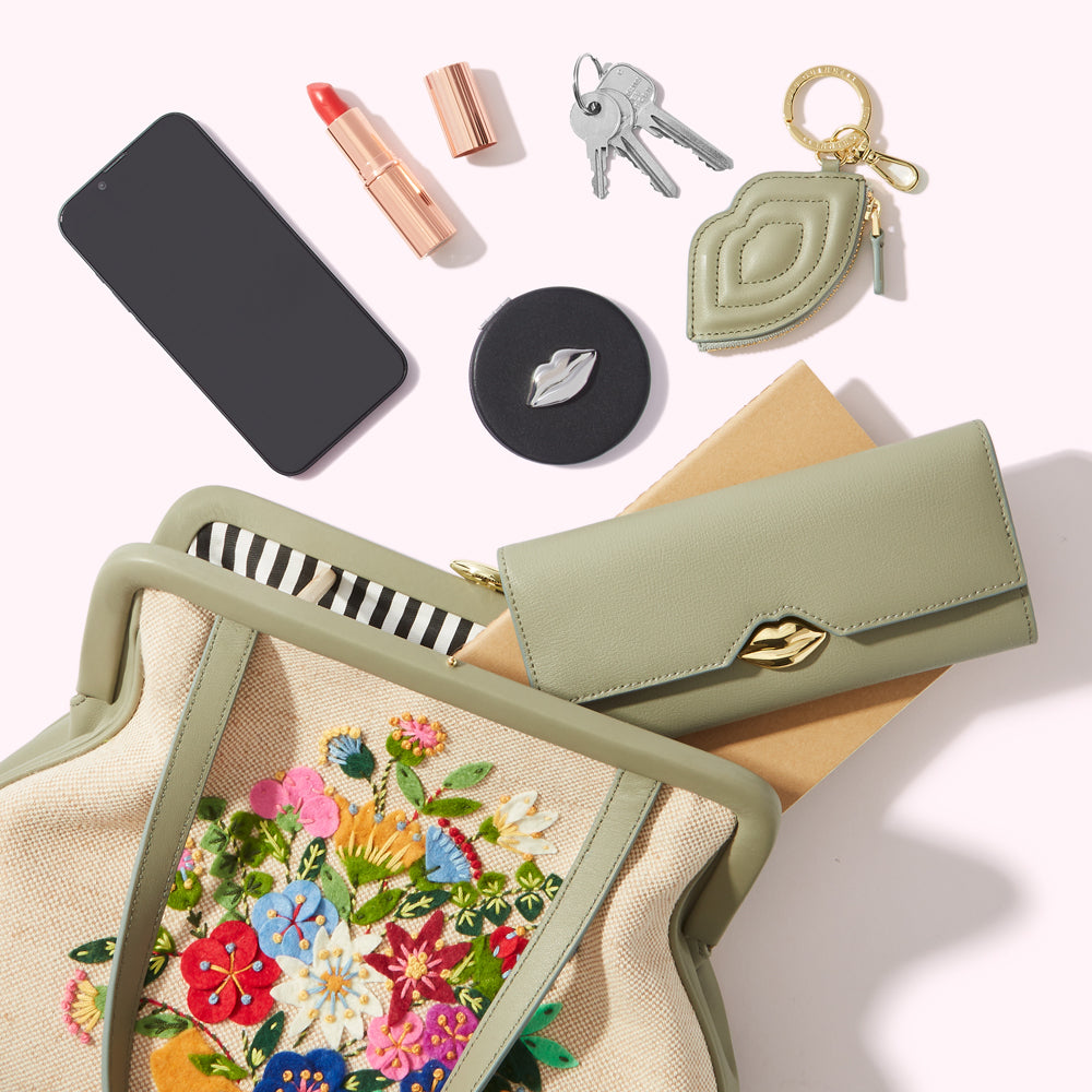 Sage green handbag with embroidered flowers, a smartphone, lipstick, keys, compact mirror, and a matching wallet on a white background.
