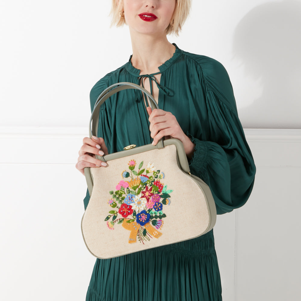 Woman in a green dress holding a beige handbag embroidered with a colourful bouquet of flowers.
