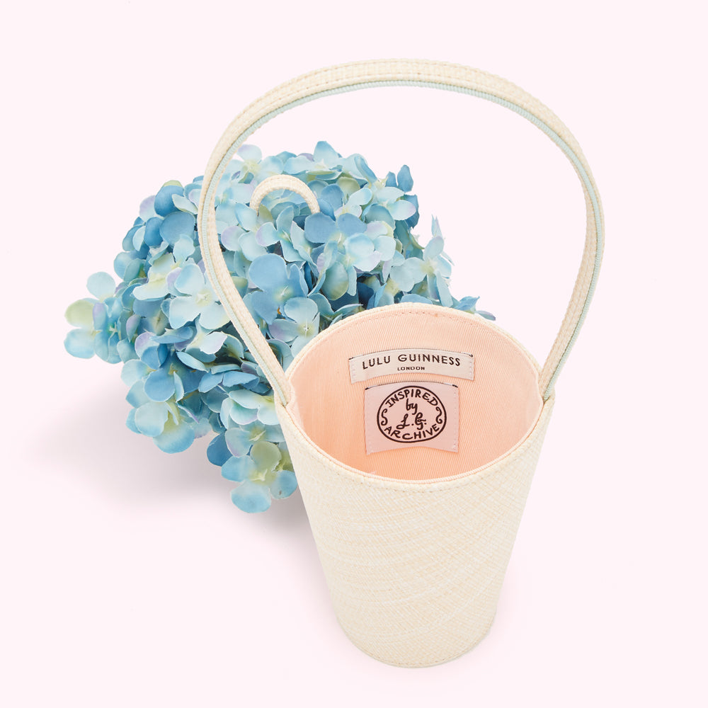 Cream-coloured Lulu Guinness bucket bag, next to a lid adorned with a bouquet of light blue hydrangeas. The bag's interior is visible and is peach-coloured, featuring the Lulu Guinness London label and an "Inspired by L.G. Archive" tag.
