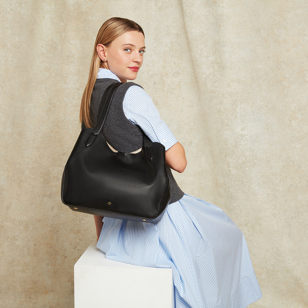Woman sitting on a plinth holding a large black leather shoulder bag against a beige background