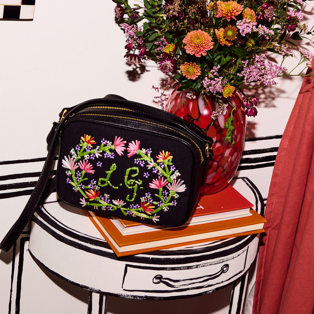 Black canvas and leather crossbody bag with wildflower embroidery on the front, embroidered L.G monogram sitting on a side table next to a vase of flowers.