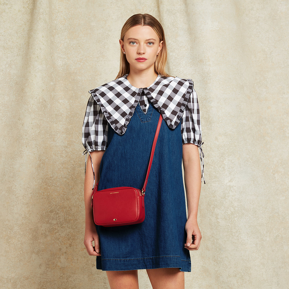Woman wearing a denim dress with a checkered blouse and carrying a red bag on a beige background