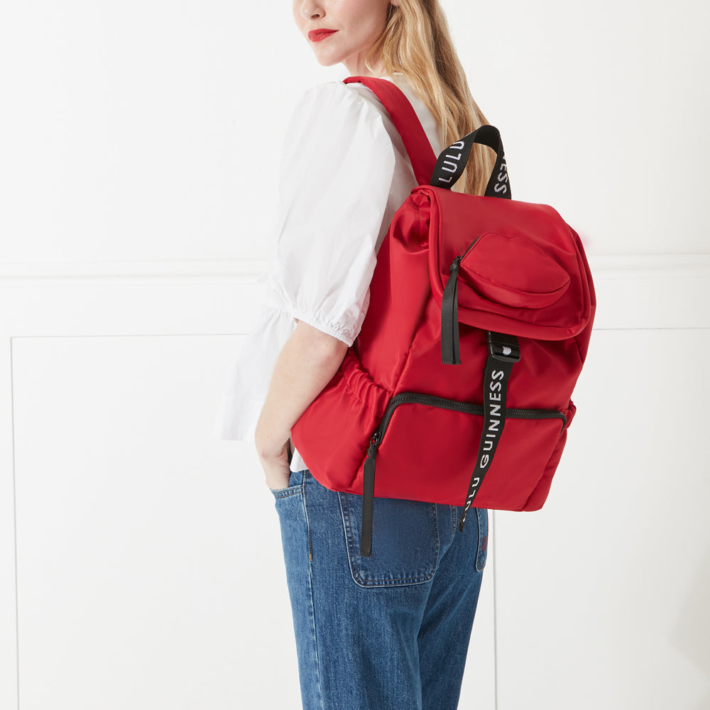 Model in a white shirt, denim dress and red lipstick wearing a red nylon shoulder bag with branded 'Lulu Guinness' shoulder strap.
