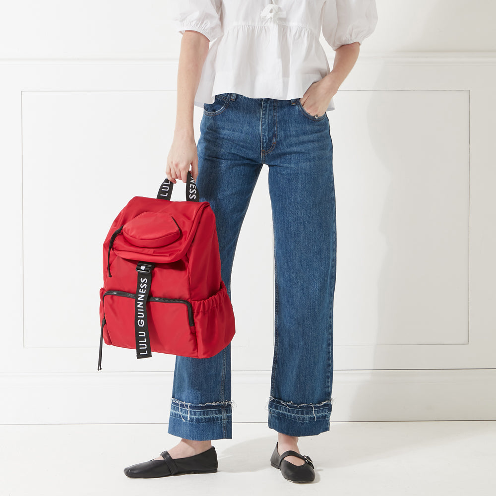 Model in a white shirt and jeans holding a red nylon backpack with 'lulu guinness' branded straps down in her hand.
