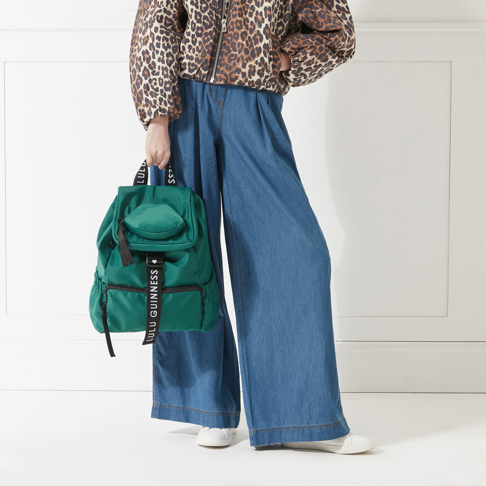 Model in a leopard print jacket and jeans holding a green nylon backpack with 'lulu guinness' branded straps down in her hand.