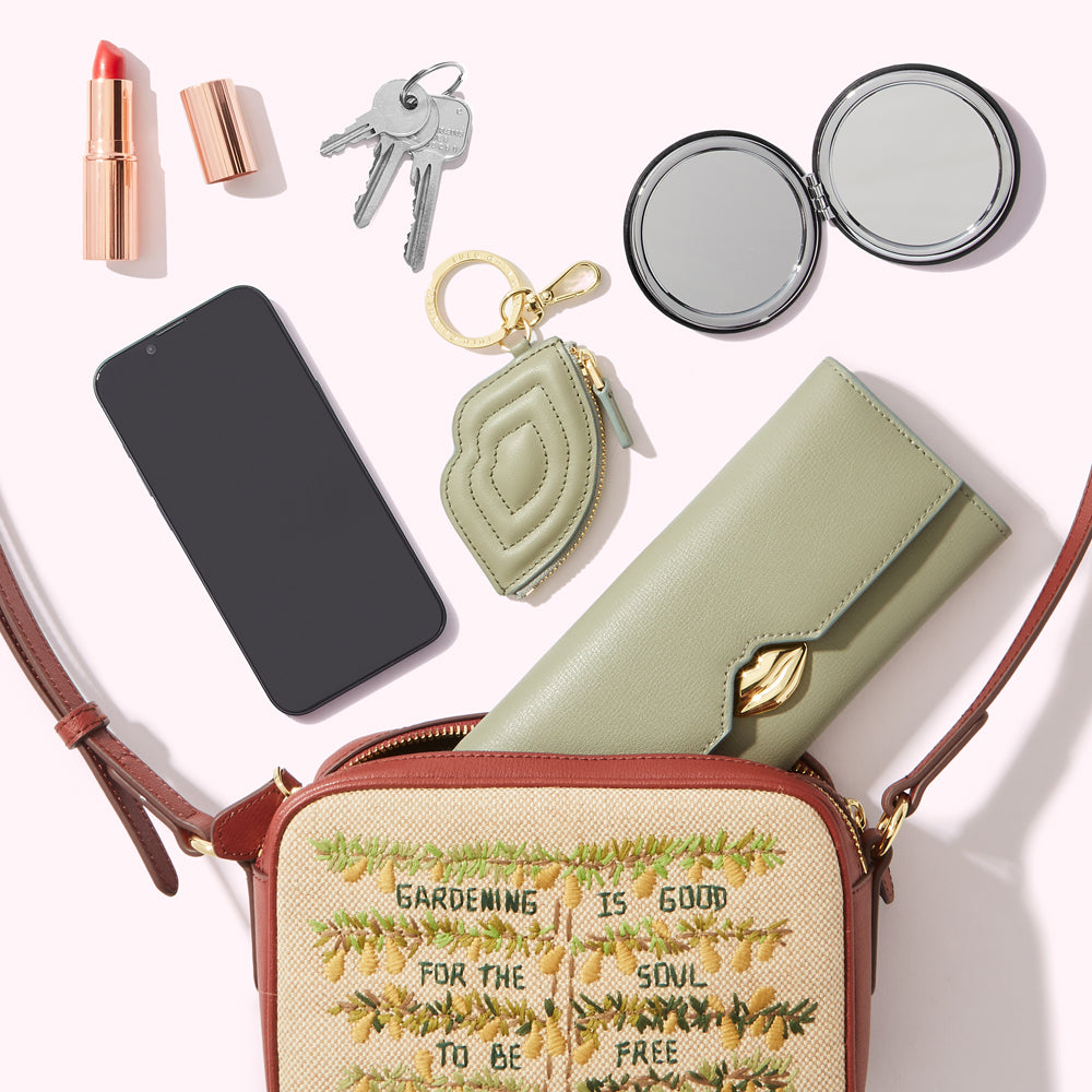 A beige crossbody bag with embroidered pears and the text "Gardening is good for the soul to be free" lies open on a white background, revealing a sage green wallet, a smartphone, keys, a compact mirror, and lipstick.
