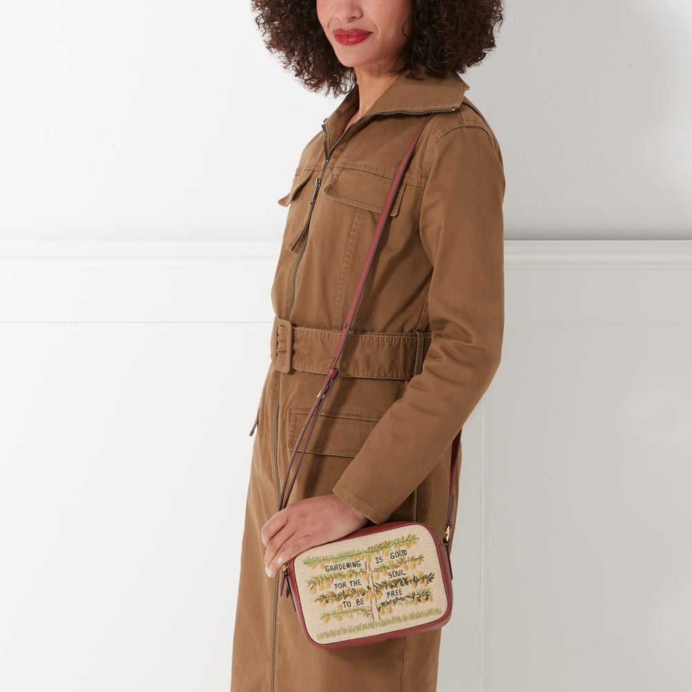 Woman in brown jacket holding a beige and brown crossbody bag with embroidered text that says "Gardening is good for the soul to be free".