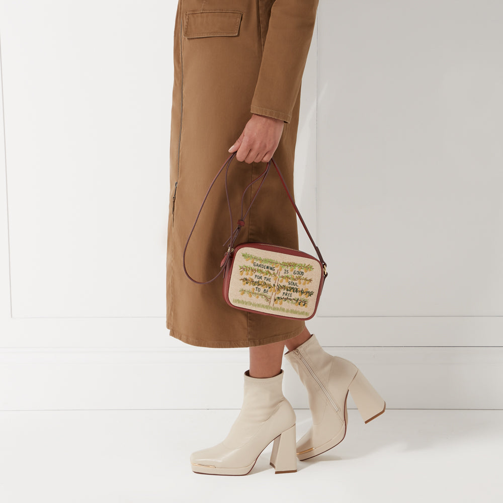 Woman in brown midi dress holding a rectangular crossbody bag with embroidered text that says "Gardening is good for the soul to be free". She's wearing cream-colored boots with thick block heels. The background is white.
