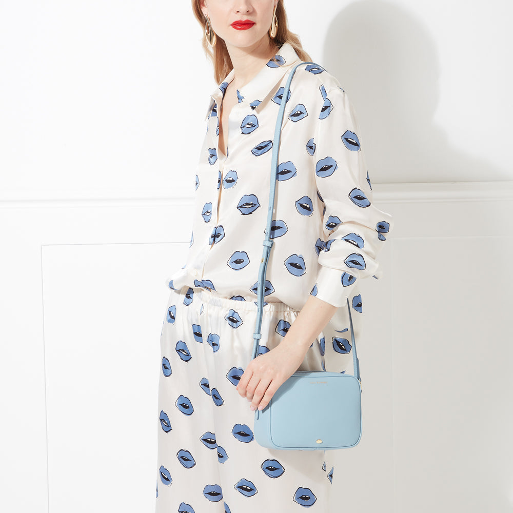 Woman in a white silk pyjama set with a blue lip print holds a pale blue crossbody bag against a white background.
