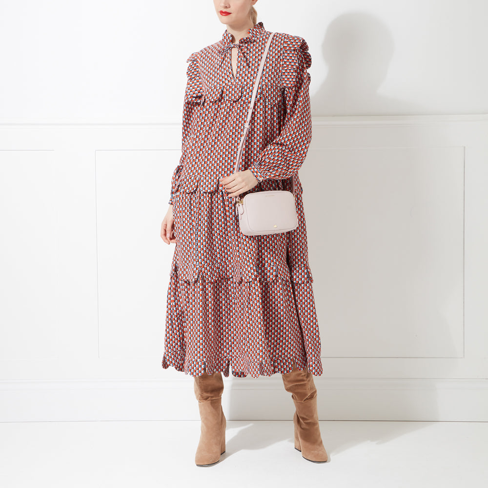 A woman in a rust-coloured midi dress and tan boots models a pale pink crossbody bag against a white background.
