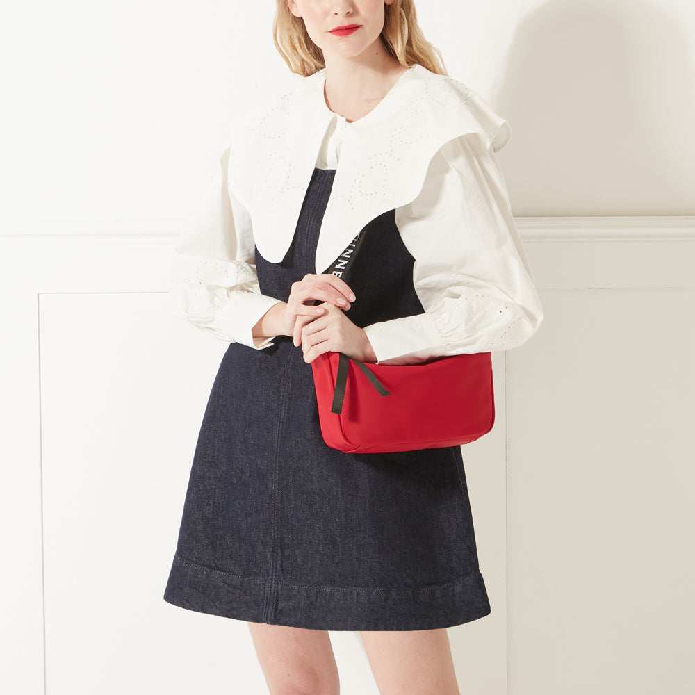 Model in a white shirt, denim dress and red lipstick wearing a red nylon shoulder bag with branded 'Lulu Guinness' shoulder strap.