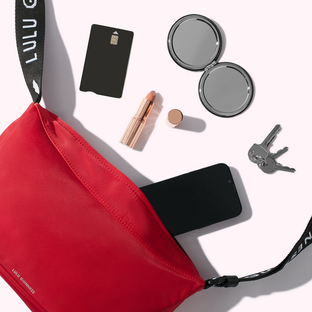 A red nylon crossbody bag with a smartphone, keys, compact mirror, card and lipstick spilling out.