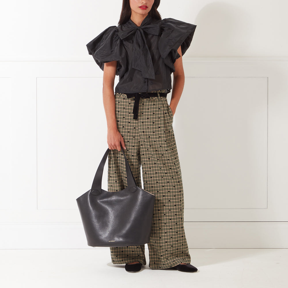 Woman wearing black puff-sleeve blouse, beige pants with black hearts, and holding a black tote bag.