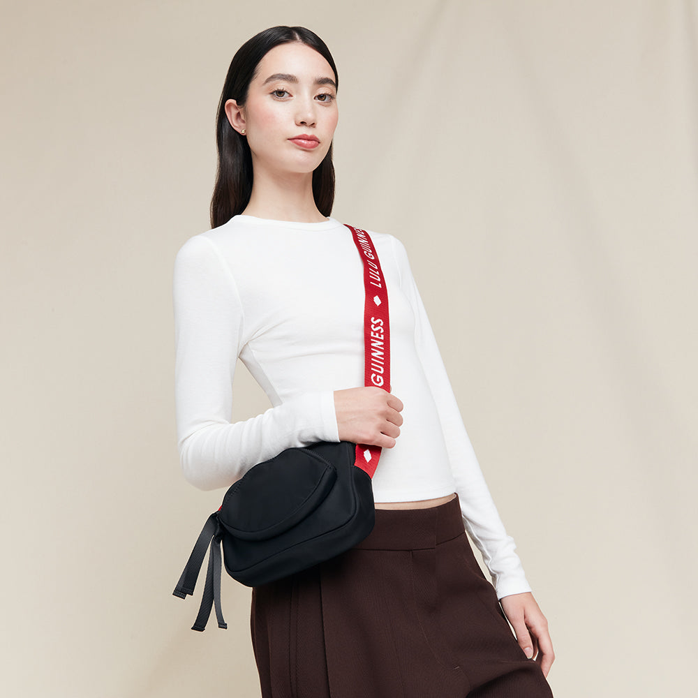 Woman holding a black bag with a red strap featuring 'Lulu Guinness' branding on a beige background