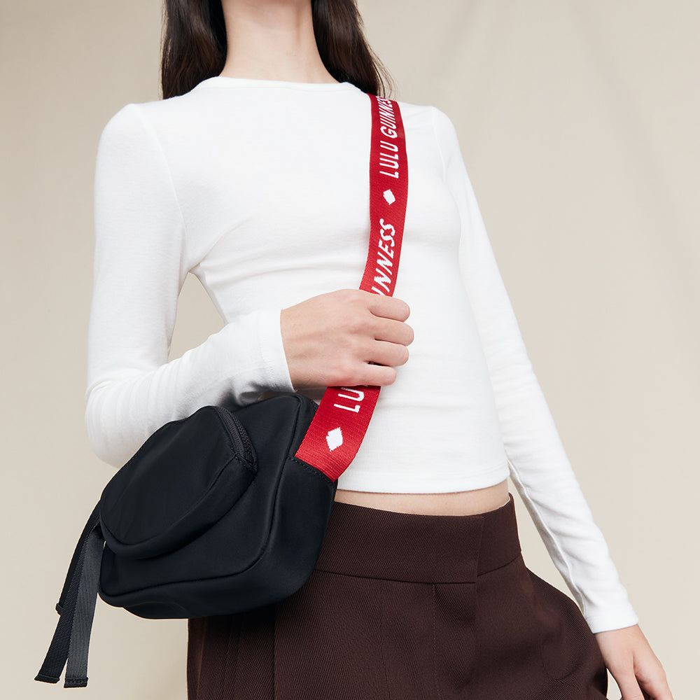 Person holding a black bag with a red strap featuring a 'Lulu Guinness' brand logo against a beige background