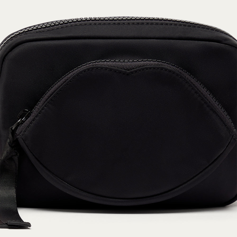 Close up image of a black crossbody bag with a lip-shaped pocket on a light grey background