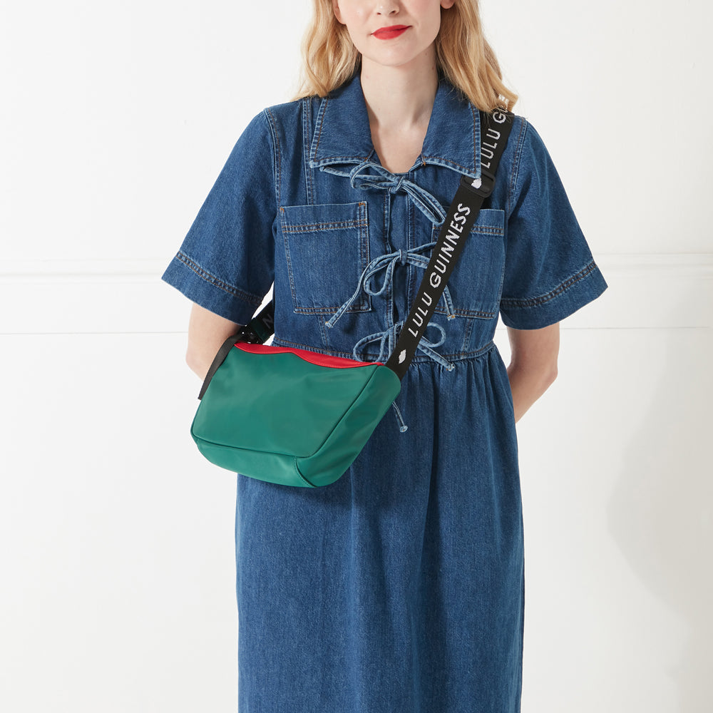 Model in a denim dress and red lipstick wearing a green and red nylon shoulder bag with branded 'Lulu Guinness' shoulder strap.