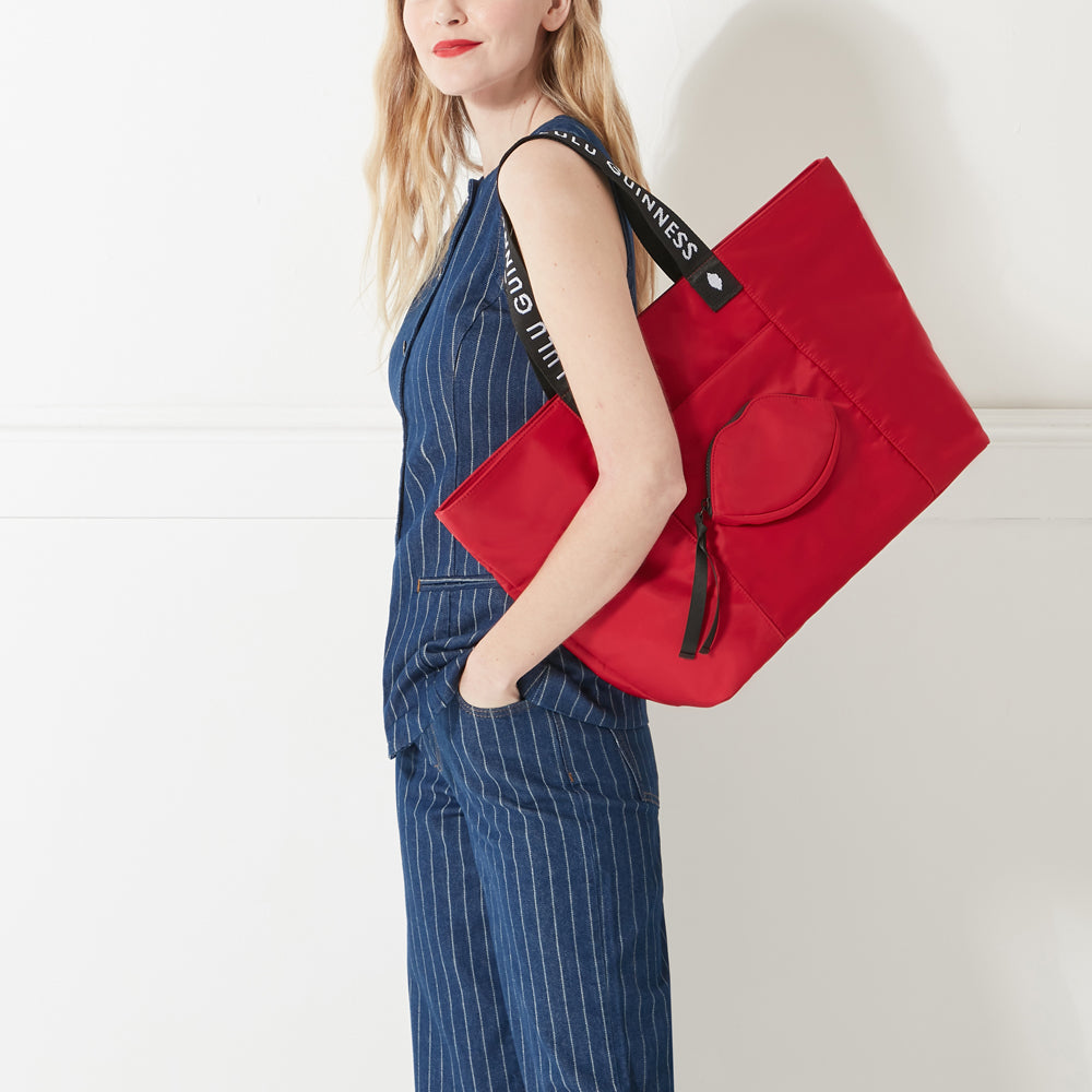 Model in denim outfit wearing a red nylon tote bag with lip shaped pocket and branded 'Lulu Guinness' straps on her arm.