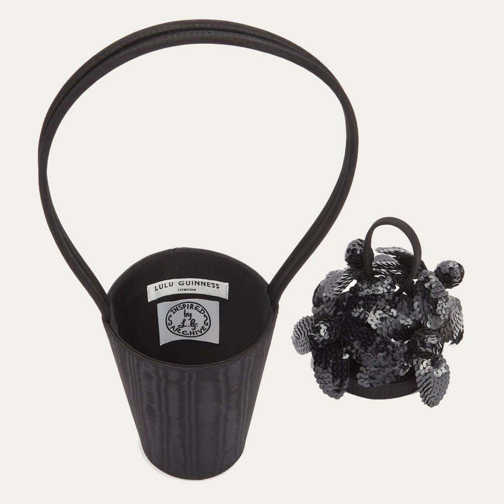 Black satin moire bucket bag with top looped handle showing inside bag revealing the branded 'Lulu Guinness London' and 'Inspired by the Archive' labels. Sitting next to the bag lid covered in black sequin flowers.