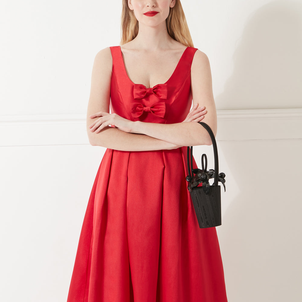 Model in red satin dress with bow details holding a black satin handbag with black sequin lily flowers on the top.