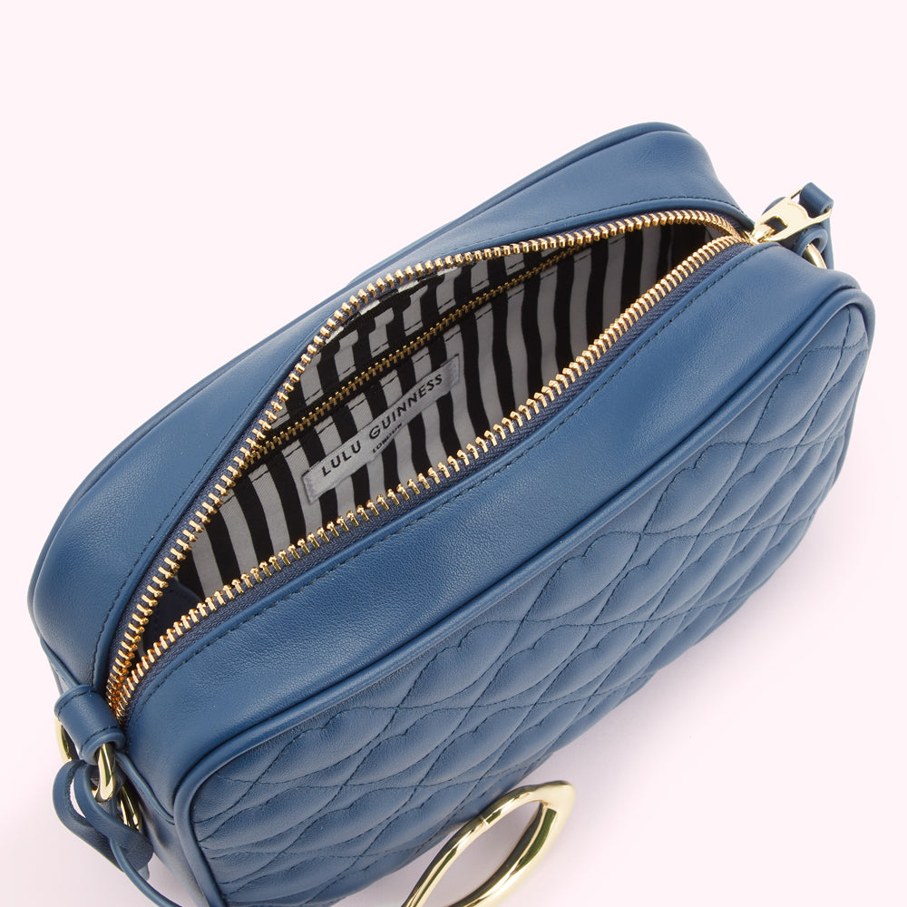 Overhead view of a blue quilted Lulu Guinness crossbody bag with gold zipper and hardware, opened to reveal its black and white striped interior.
