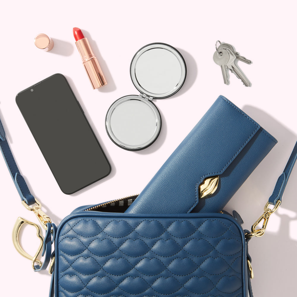 A blue quilted crossbody bag with a gold clasp, containing a blue wallet, a smartphone, a compact mirror, lipstick, and keys, all on a white background.
