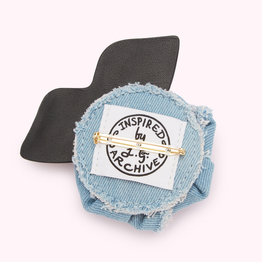 Back view of a light blue denim flower brooch with black leather accents and a gold pin. The back features a label that reads "Inspired by J.G. Archive".
