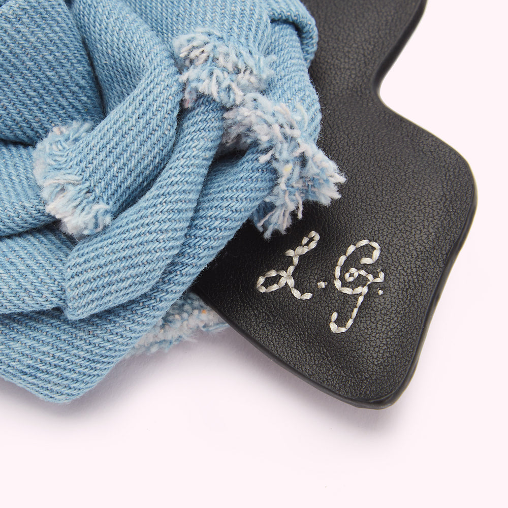Close-up of a light blue denim knotted hair clip with "L.G." embroidered on a black leather base against a white background.
